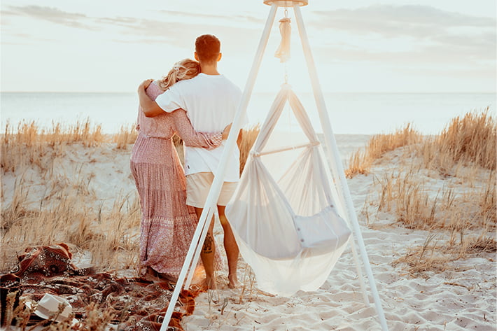 On the beach, a couple hug each other and look into the sunset. In front of them is a NONOMO with a tepee frame. 